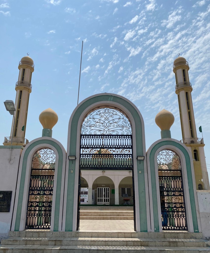 Picture of Nabih Saleh shrine from its entrance. A simple arching gate with greens and whites. Two minarets in the background. Within, a tiled courtyard and an inner shrine.